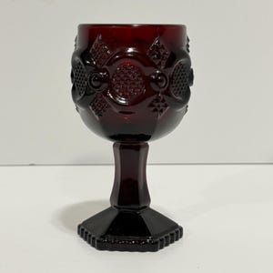 May include: A deep red glass goblet featuring a textured pattern of raised circles and diamond shapes on the bowl. The stem is faceted and leads to a hexagonal base, creating a structured silhouette for this pressed glass drinking vessel.