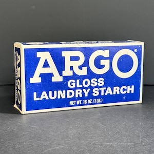 May include: A rectangular box of Argo Gloss Laundry Starch with a blue background and white text. The front label displays the brand name ARGO in large letters, followed by GLOSS LAUNDRY STARCH and the net weight of 16 oz. (1 lb.) in white.