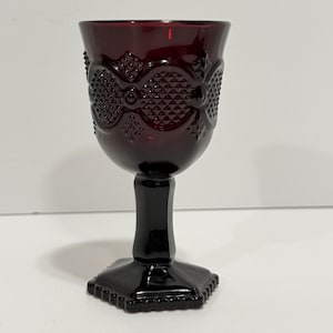 May include: A deep red glass goblet featuring a textured diamond pattern on the bowl. The stem is smooth and cylindrical, leading down to a hexagonal base with a beaded edge. The glass is translucent, allowing light to pass through the dark red color.