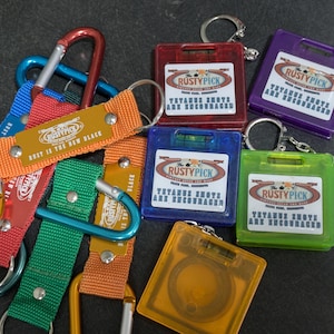 May include: A collection of keychains and carabiners in various colors. The keychains are rectangular with the text "RUSTYPICK" and "YAYABUSA ARE ENCOURAGED". The carabiners are red, blue, green, and gold, with orange and blue straps that read "RUST IS THE NEW BLACK".