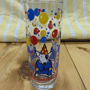 May include: A clear glass with a colorful design featuring a white bull terrier wearing a blue sweater and a party hat. The glass has red, yellow, and blue balloons and confetti. The text on the glass reads "Buds Mackenzy's Original Party Animal".