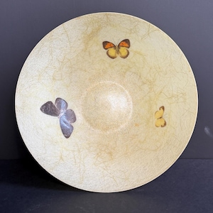 May include: A round, shallow bowl with a cream-colored, fibrous, textured surface. The interior features three distinct butterfly illustrations: one orange and yellow butterfly at the top, one dark grey butterfly on the left, and a small yellow butterfly on the right.