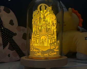 May include: A glass dome with a wooden base, containing a paper cut-out design of a castle with a Ferris wheel and a carousel. The design is illuminated from within, casting a warm glow. The base is inscribed with the names "Lyubc and Luynh".