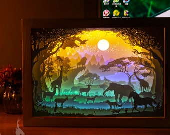 Könnte beinhalten: Ein 3D-Papier-Schnittbild, das eine afrikanische Landschaft mit einem leuchtenden Mond und Sternen darstellt. Die Szene umfasst Bäume, Berge und Tiere wie Giraffen, Elefanten und Zebras. Das Kunstwerk ist in einem hellen Holzrahmen gerahmt und misst 270 x 200 mm.