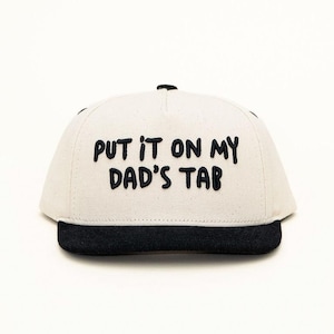 May include: A baseball cap with a cream-coloured crown and a black brim. The text "PUT IT ON MY DAD'S TAB" is printed in black on the front of the cap. The cap is set against a white background.