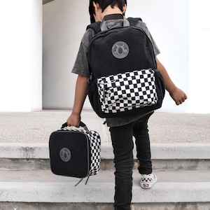 以下が含まれることがあります： 黒のコーデュロイのバックパックに、チェック柄のポケットと黒いランチバッグが付いています。バックパックには、「Big Top Co.」の文字と松の木のデザインが描かれた丸いパッチが付いています。