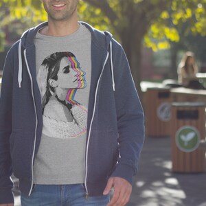 May include: A gray t-shirt with a graphic print of a woman's face in a layered, colorful design. The woman's face is shown in a series of overlapping images, each with a different color overlay. The colors used are pink, yellow, blue, and green.