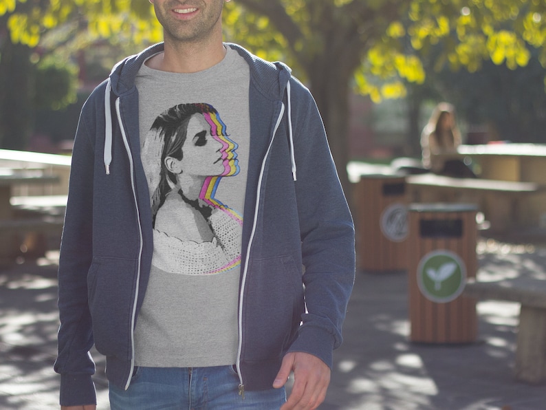 May include: A gray t-shirt with a graphic print of a woman's face in a layered, colorful design. The woman's face is shown in a series of overlapping images, each with a different color overlay. The colors used are pink, yellow, blue, and green.