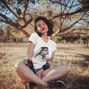 May include: A woman wearing a white t-shirt with a colorful graphic of a woman's face. She is sitting in a field of tall grass.