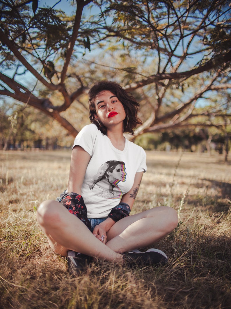 May include: A woman wearing a white t-shirt with a colorful graphic of a woman's face. She is sitting in a field of tall grass.