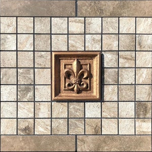 May include: A gold-colored fleur-de-lis tile accent in a brown frame, set against a background of small, square, beige tiles.