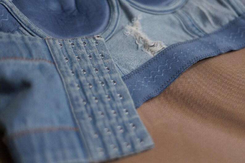 May include: A close-up of a blue denim bra with a hook and eye closure. The bra has a decorative stitching detail along the edge.