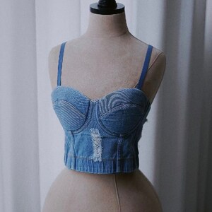 May include: A blue denim corset top with adjustable straps. The top has a sweetheart neckline and is made of a distressed denim fabric.