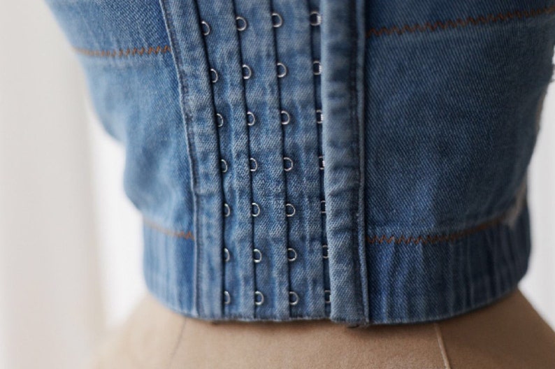 May include: A close-up of a denim corset with a row of metal eyelets and a brown stitching detail.
