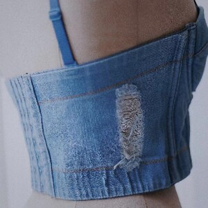 May include: A blue denim corset with a distressed detail on the front. The corset has a boned structure and adjustable straps.