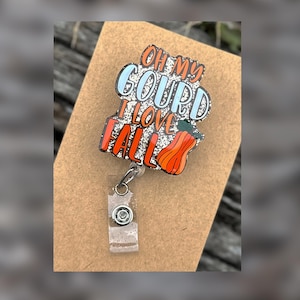 May include: A badge reel featuring a decorative acrylic topper with a glittery white background and the orange and blue text "OH MY GOURD I LOVE FALL" accompanied by a small orange pumpkin illustration with a green stem and a clear plastic strap.