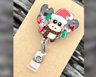 Può includere: Un portabadge con un personaggio a forma di alce marrone glitterato che indossa un cappello da Babbo Natale rosso e bianco e una sciarpa verde a pois rossi. L'alce è circondato da lampadine natalizie rosse e verdi. Il portabadge ha un cinturino.