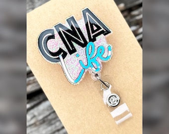 May include: A badge reel featuring a glittery acrylic topper with the black text CNA and the light blue cursive text life. The topper has a pink and white iridescent glitter background and is attached to a clear plastic strap with a metal snap.