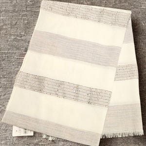 May include: A folded scarf with a cream-colored background and horizontal gray stripes. The stripes are accented with small, shimmering embellishments. The scarf has a fringed edge and a tag in the corner.