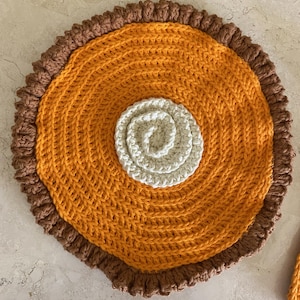 May include: A handmade crochet placemat resembling a pumpkin pie. The pie features a golden-orange center, a light beige swirl in the middle, and a brown, ruffled crust. The circular design is perfect for adding a touch of warmth to any table setting.