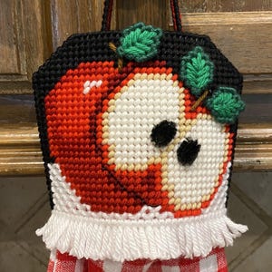 May include: A handmade kitchen towel holder with a cross-stitched apple design, green leaves, and a black background. It includes a red and white checked towel and a white fringe.