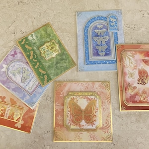 Op de afbeelding: Een verzameling decoratieve wenskaarten met vlinderontwerpen. De kaarten hebben verschillende kleuren, waaronder oranje, groen, blauw en paars, met gouden folie accenten. Sommige kaarten hebben de tekst "Beautiful" en "To Say".