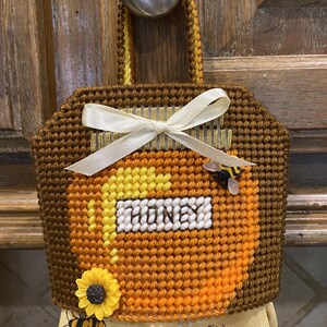 Puede incluir: Un portatoallas de cocina hecho a mano, marrón y amarillo, con un diseño de tarro de miel. La palabra "HONEY" se muestra en la parte delantera. Un girasol amarillo y una pequeña abeja están unidos al soporte. Una cinta blanca está atada en la parte superior.
