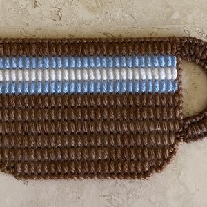 Pu&ograve; includere: Una tazza da caff&egrave; in crochet marrone con un motivo a righe blu e bianche.