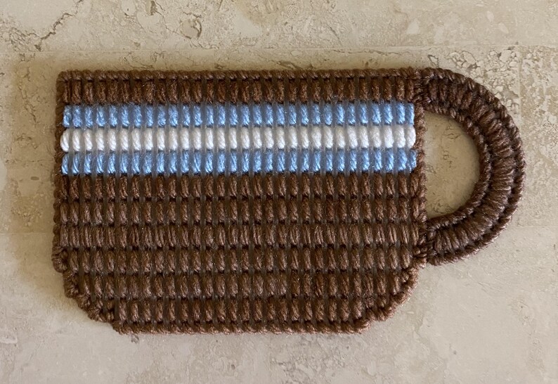 Pu&ograve; includere: Una tazza da caff&egrave; in crochet marrone con un motivo a righe blu e bianche.