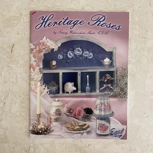 May include: A book cover titled "Heritage Roses" featuring a painted shelf with decorative items. The shelf displays a clock, a piggy bank, a bell, and a vase with flowers. A teapot, teacup, and cookies are on a table.