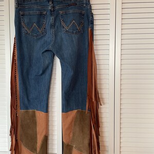 May include: Blue denim jeans with brown suede patchwork on the lower legs and long brown fringe detailing along the sides. The jeans have the Wrangler logo on the back pockets and are hanging on a hanger.