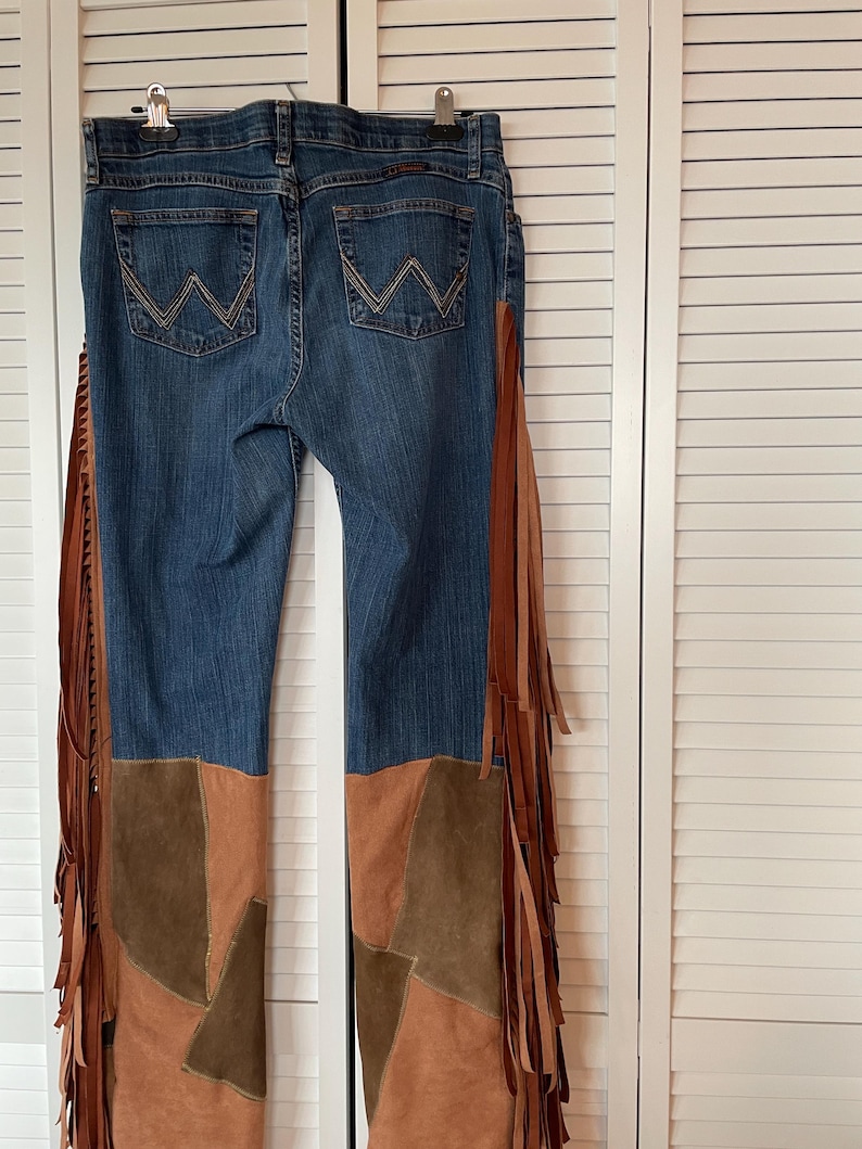May include: Blue denim jeans with brown suede patchwork on the lower legs and long brown fringe detailing along the sides. The jeans have the Wrangler logo on the back pockets and are hanging on a hanger.