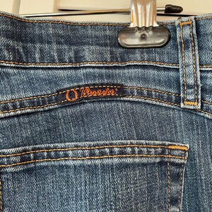 May include: Close-up of blue denim jeans with orange stitching. The Wrangler logo is visible on the back pocket. The jeans are hanging from a silver hanger.