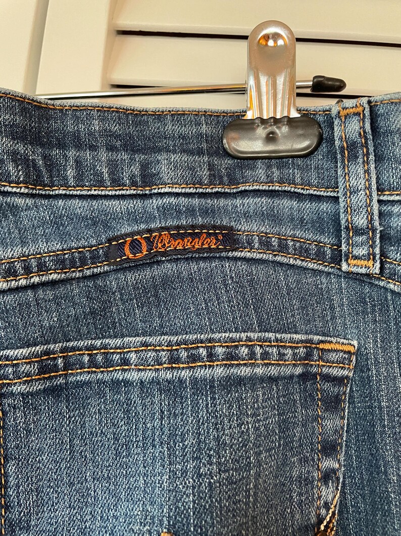 May include: Close-up of blue denim jeans with orange stitching. The Wrangler logo is visible on the back pocket. The jeans are hanging from a silver hanger.