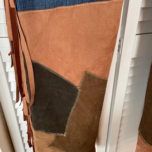 May include: Blue denim jeans with brown suede patchwork and fringe detailing. The patchwork includes various shades of brown, creating a unique and textured design. The fringe adds a bohemian flair to the jeans.