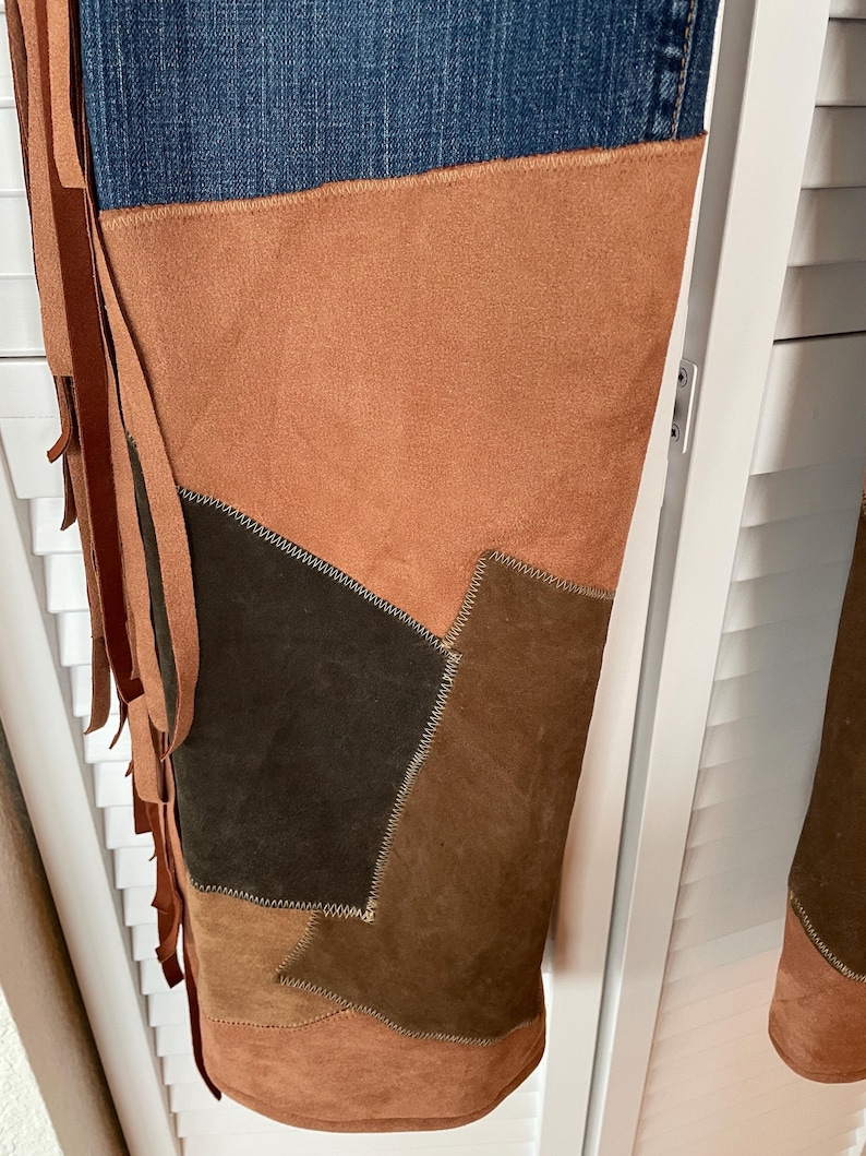 May include: Blue denim jeans with brown suede patchwork and fringe detailing. The patchwork includes various shades of brown, creating a unique and textured design. The fringe adds a bohemian flair to the jeans.