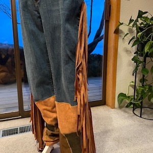 May include: Blue denim jeans with brown suede patches and long fringe detailing along the sides. The jeans have a unique, bohemian style, with a patchwork design on the lower legs.