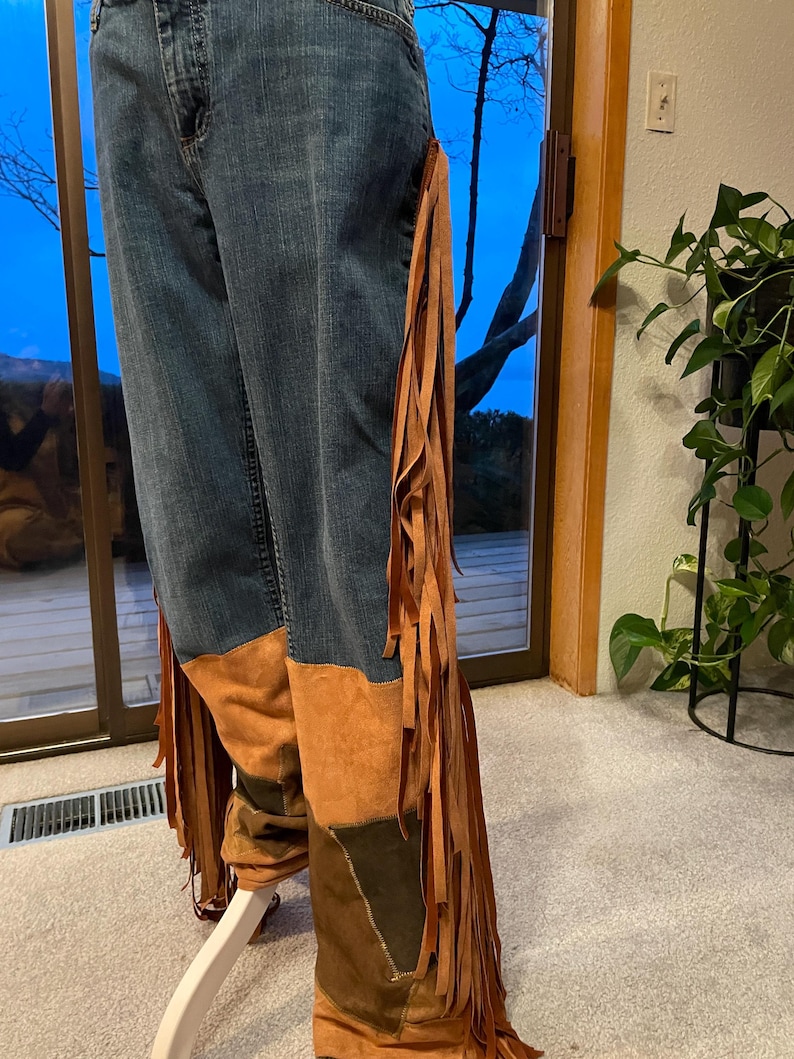 May include: Blue denim jeans with brown suede patches and long fringe detailing along the sides. The jeans have a unique, bohemian style, with a patchwork design on the lower legs.