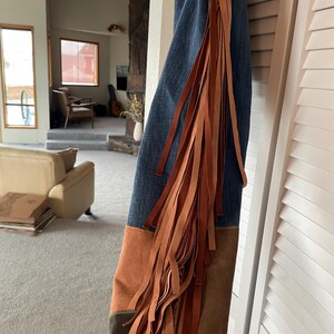 May include: Blue denim jeans with brown suede patches and long, flowing brown fringe along the side. The jeans are hanging against a white door. The style is reminiscent of a Western or bohemian aesthetic.