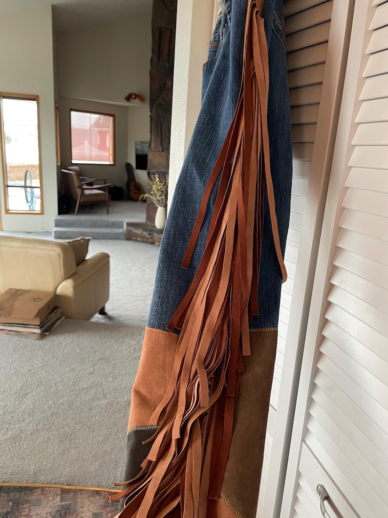 May include: Blue denim jeans with brown suede patches and long, flowing brown fringe along the side. The jeans are hanging against a white door. The style is reminiscent of a Western or bohemian aesthetic.