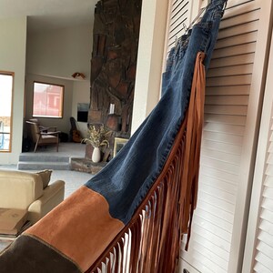 May include: Blue denim jeans with brown suede and fringe detailing. The jeans have a dark wash and feature a suede panel on the side with long fringe. The jeans are hanging on a white door.