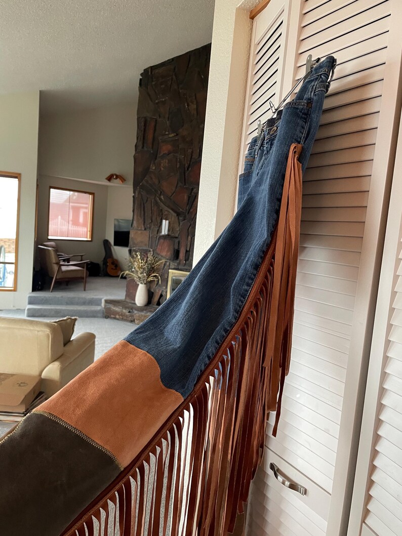 May include: Blue denim jeans with brown suede and fringe detailing. The jeans have a dark wash and feature a suede panel on the side with long fringe. The jeans are hanging on a white door.