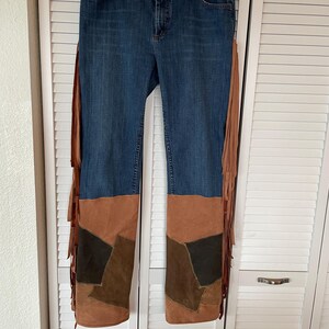 May include: Blue denim jeans with brown suede patchwork on the lower legs and long fringe detailing on the sides. The jeans are hanging on a hanger against a white background.