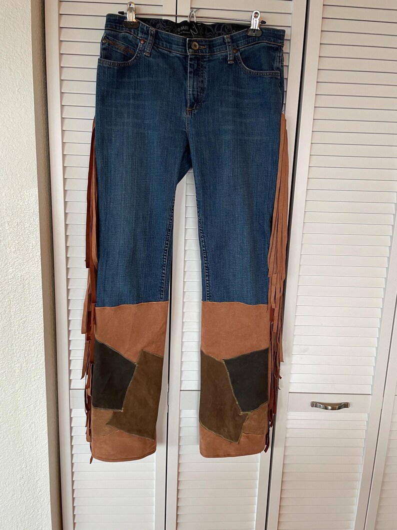 May include: Blue denim jeans with brown suede patchwork on the lower legs and long fringe detailing on the sides. The jeans are hanging on a hanger against a white background.