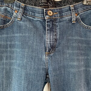 May include: Close-up of blue denim jeans with a button closure and a decorative waistband. The jeans have classic five-pocket styling and are hanging from a metal hanger. The brand name "Q-BABY" is visible on the waistband.