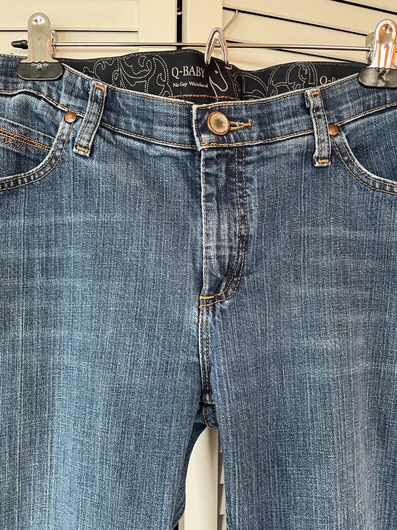 May include: Close-up of blue denim jeans with a button closure and a decorative waistband. The jeans have classic five-pocket styling and are hanging from a metal hanger. The brand name "Q-BABY" is visible on the waistband.