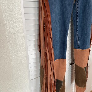 May include: Blue denim pants with brown suede fringe and patchwork details. The fringe runs along the outer seams, and the lower legs feature brown suede patches in various shades.