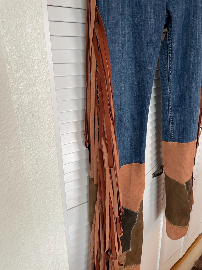May include: Blue denim pants with brown suede fringe and patchwork details. The fringe runs along the outer seams, and the lower legs feature brown suede patches in various shades.