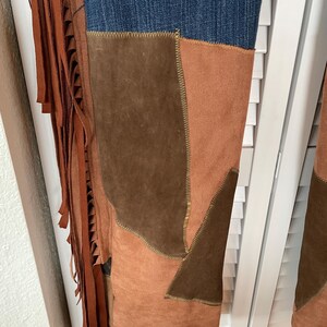 May include: Close-up of denim jeans with suede patchwork in shades of brown and tan. The jeans feature a fringed leather detail on the side, adding a bohemian flair. The stitching is visible, highlighting the craftsmanship of the garment.