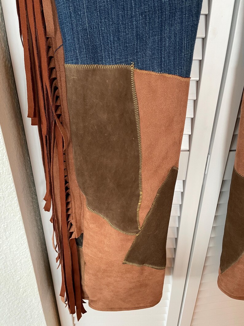 May include: Close-up of denim jeans with suede patchwork in shades of brown and tan. The jeans feature a fringed leather detail on the side, adding a bohemian flair. The stitching is visible, highlighting the craftsmanship of the garment.